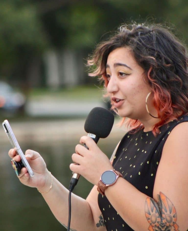 AP speaks truth into the mic. AP the Poet has wavy mid length ombre hair, hoop earrings, a colorful tattoo and a loving look out into the crowd. The water of the Loose Park pond is reflecting the trees behind her as AP shares her poem.
